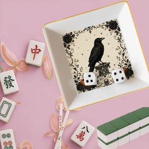 May include: A square white ceramic trinket dish with a gold rim featuring a black raven illustration, floral accents, and a skull. Two white six-sided dice rest inside. Scattered around are various mahjong tiles and light pink flower petals.