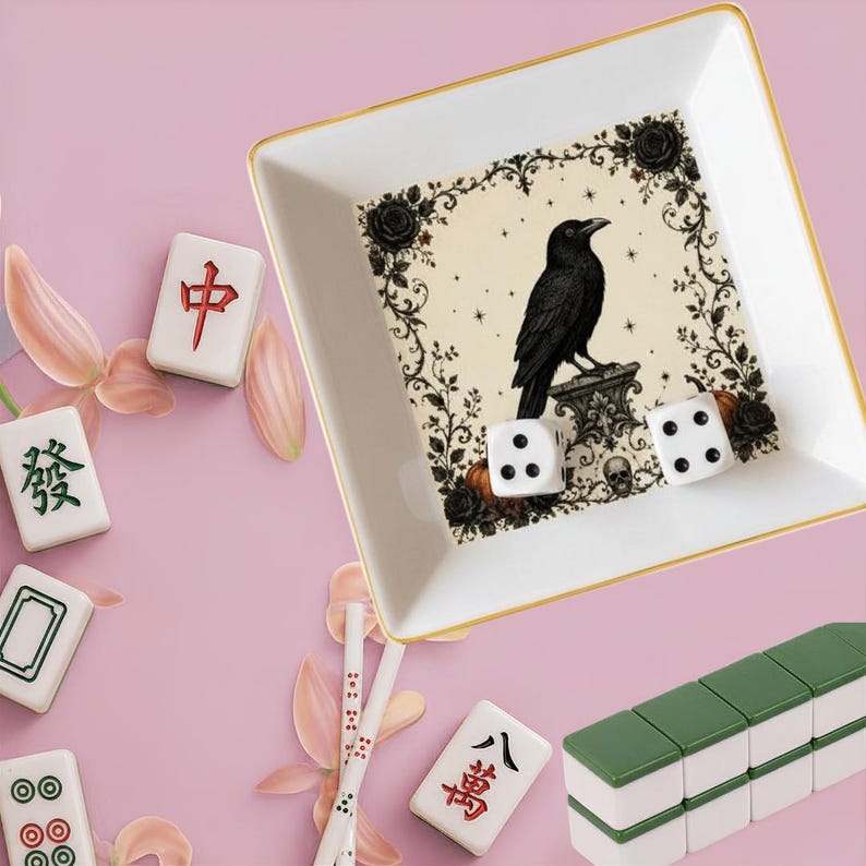 May include: A square white ceramic trinket dish with a gold rim featuring a black raven illustration, floral accents, and a skull. Two white six-sided dice rest inside. Scattered around are various mahjong tiles and light pink flower petals.
