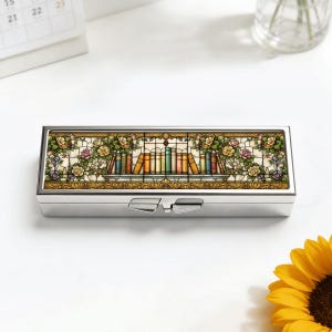 May include: A rectangular, silver pill box with a stained-glass design. The design features a row of colorful books on a shelf, surrounded by roses and foliage. The box has a hinged lid and a clasp closure.