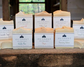 May include: An assortment of goat milk soaps with white labels featuring a forest logo and the brand name "OUTPOST". The soaps are various shades of beige and cream, with scents like Lavender Sage and Winter Pine.