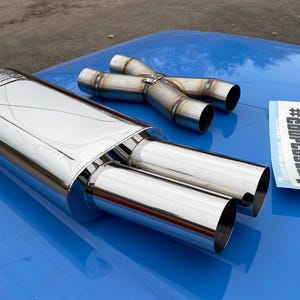Pode incluir: Um silencioso de escape cromado polido com saídas duplas, um tubo em Y e um autocolante que diz "#COUPCUSGUM" numa superfície azul. O silencioso tem um corpo retangular e dois tubos de escape redondos.