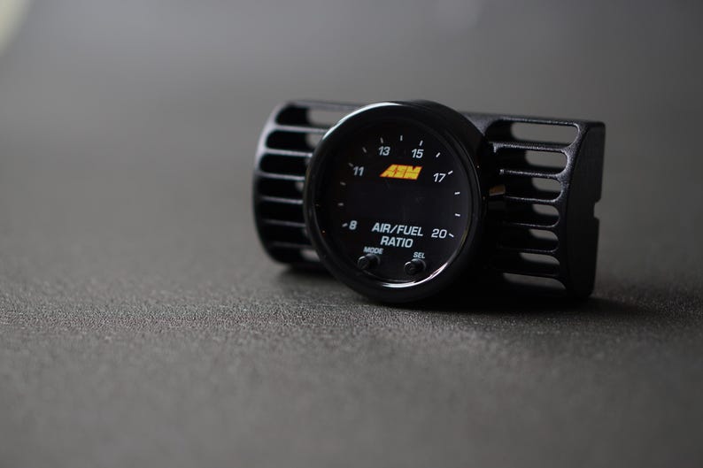 Puede incluir: Medidor de relaci&oacute;n aire/combustible negro con una carcasa negra. El medidor muestra n&uacute;meros del 8 al 20 y el texto "AIR/FUEL RATIO". El medidor est&aacute; montado en una carcasa negra similar a una rejilla de ventilaci&oacute;n.