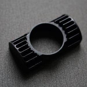 Puede incluir: Un anillo negro con un dise&ntilde;o rectangular con ranuras verticales en cada lado y una abertura circular en el centro. El anillo est&aacute; hecho de un material negro mate y se encuentra sobre una superficie gris oscuro.