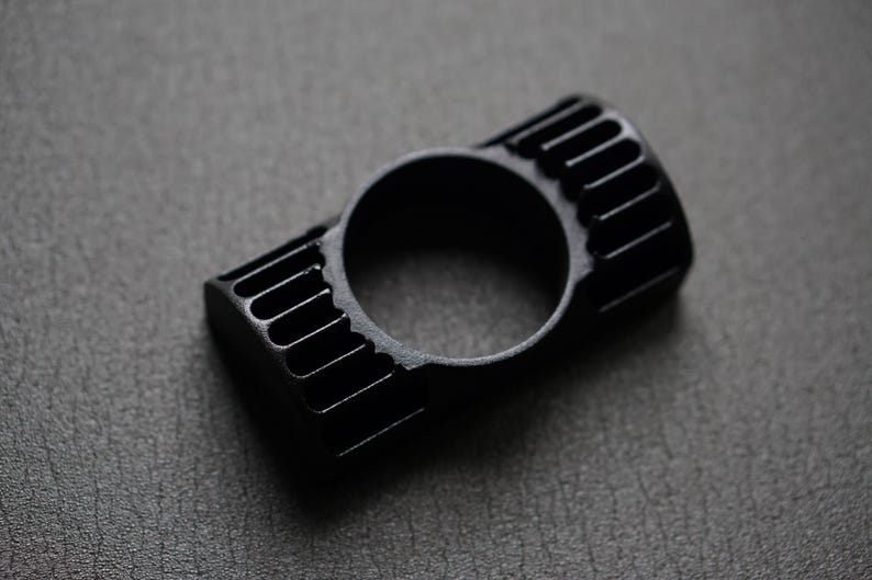 Puede incluir: Un anillo negro con un dise&ntilde;o rectangular con ranuras verticales en cada lado y una abertura circular en el centro. El anillo est&aacute; hecho de un material negro mate y se encuentra sobre una superficie gris oscuro.