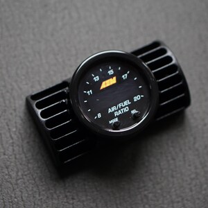 Puede incluir: Un medidor de relaci&oacute;n aire/combustible negro con marcas blancas y el texto "AIR/FUEL RATIO 20-". El medidor est&aacute; montado sobre una base negra ranurada. La esfera del medidor muestra n&uacute;meros y el logotipo de AEM.