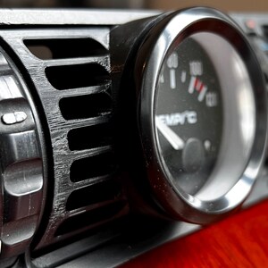 Puede incluir: Primer plano de una rejilla de ventilaci&oacute;n negra del salpicadero de un coche con un indicador adjunto. El indicador tiene un bisel plateado y una esfera negra con marcas blancas y una aguja roja. La rejilla tiene una serie de lamas horizontales.