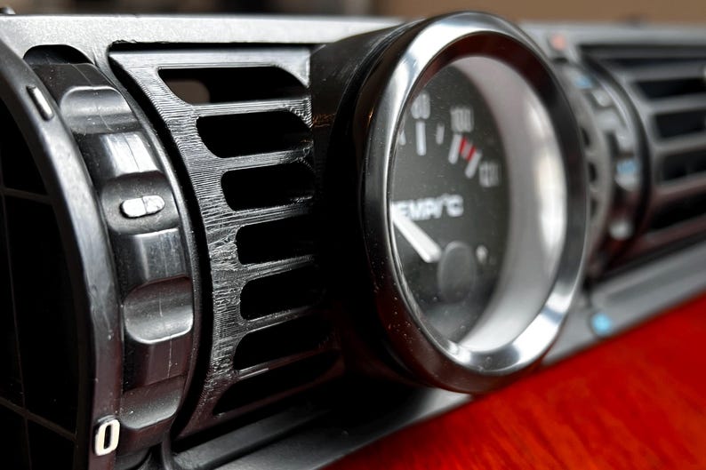 Puede incluir: Primer plano de una rejilla de ventilaci&oacute;n negra del salpicadero de un coche con un indicador adjunto. El indicador tiene un bisel plateado y una esfera negra con marcas blancas y una aguja roja. La rejilla tiene una serie de lamas horizontales.