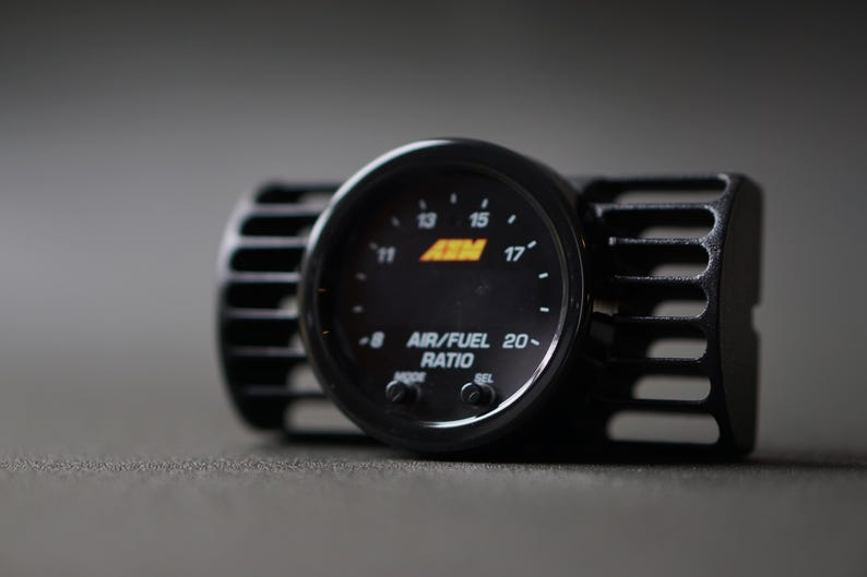 Puede incluir: Un medidor de relaci&oacute;n aire/combustible negro con una pantalla digital, con el texto "AIR/FUEL RATIO" y el logotipo de la marca "AEM". El medidor est&aacute; montado en una carcasa negra con un dise&ntilde;o ranurado.