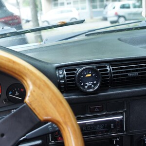 Puede incluir: Vista interior del salpicadero de un coche cl&aacute;sico. El volante de madera est&aacute; en primer plano, con indicadores y un salpicadero negro. Un indicador redondo est&aacute; montado en la rejilla de ventilaci&oacute;n central.
