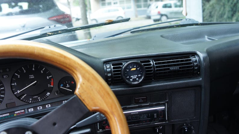 Puede incluir: Vista interior del salpicadero de un coche cl&aacute;sico. El volante de madera est&aacute; en primer plano, con indicadores y un salpicadero negro. Un indicador redondo est&aacute; montado en la rejilla de ventilaci&oacute;n central.