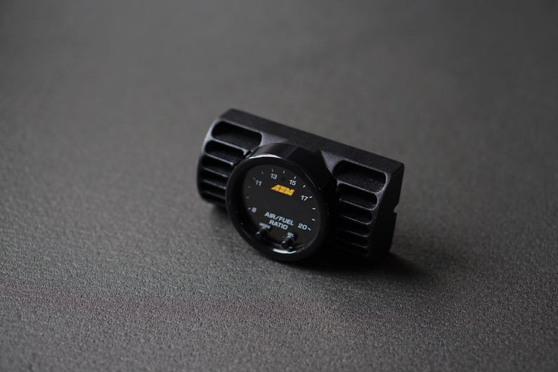 Puede incluir: Medidor de relaci&oacute;n aire/combustible negro con pantalla digital. El medidor tiene una carcasa negra con ranuras verticales en los lados. La esfera muestra n&uacute;meros y el texto "AIR/FUEL RATIO".