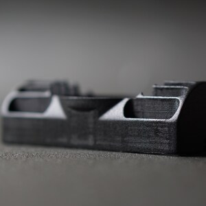 Puede incluir: Un objeto rectangular gris oscuro con una superficie texturizada. El objeto tiene una serie de formas recortadas y un extremo redondeado. Probablemente sea un componente o parte de un dispositivo m&aacute;s grande.