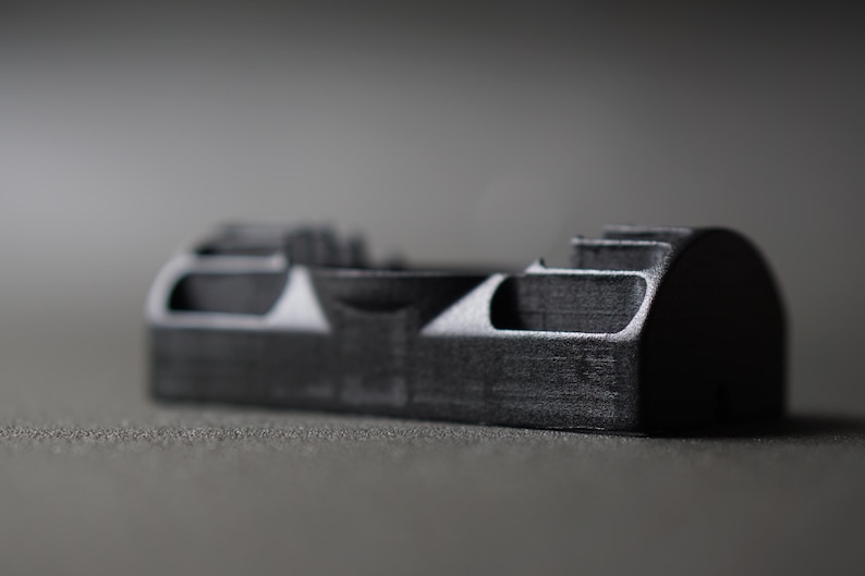 Puede incluir: Un objeto rectangular gris oscuro con una superficie texturizada. El objeto tiene una serie de formas recortadas y un extremo redondeado. Probablemente sea un componente o parte de un dispositivo m&aacute;s grande.