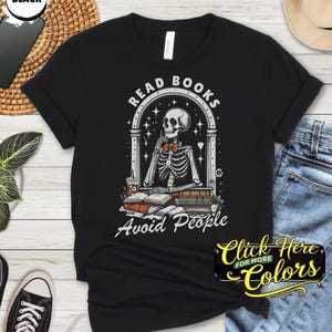 May include: Black t-shirt with a graphic of a skeleton reading books, with the text "Read Books Avoid People". The skeleton is in a window frame, with books and a coffee cup. The shirt is a classic crew neck.