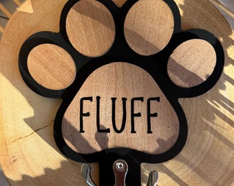 May include: Wooden paw print-shaped wall hook with the word "FLUFF" in black lettering. The paw print has a black outline and a natural wood grain finish. A silver-colored double hook is attached below the paw print.