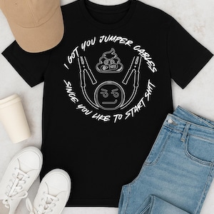 May include: Black t-shirt with a white graphic that includes the text "I GOT YOU JUMPER CABLES SINCE YOU LIKE TO START SHIT". The graphic also features jumper cables, a poop emoji, and a frowning face. A tan baseball cap, sunglasses, white sneakers, and blue jeans are also in the image.