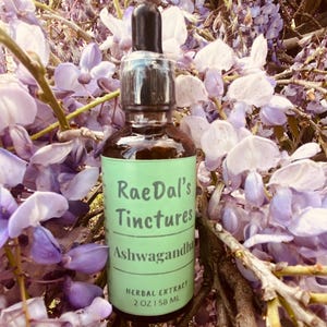 May include: A glass bottle with a dropper top and a green label that reads "RaeDal's Tinctures Ashwagandha Herbal Extract 2 oz | 58 ML". The bottle is sitting on a bed of purple flowers.