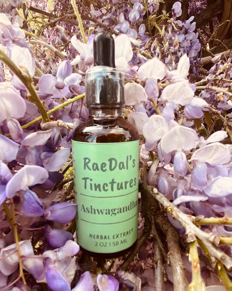 May include: A glass bottle with a dropper top and a green label that reads "RaeDal's Tinctures Ashwagandha Herbal Extract 2 oz | 58 ML". The bottle is sitting on a bed of purple flowers.