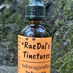 May include: A brown glass bottle of Rae Dal's Tinctures Ashwagandha for dogs or cats. The bottle has a black dropper and an orange label with white text. The label reads "Herbal Extract 2 OZ | 58 ML".