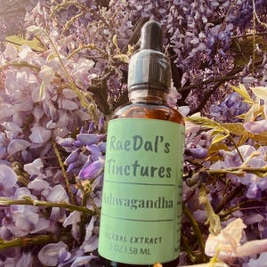 May include: A green glass bottle with a dropper top and a label that reads "RaeDal's Tinctures Ashwagandha Herbal Extract 2 oz | 58 ML". The bottle is sitting on a bed of purple flowers.