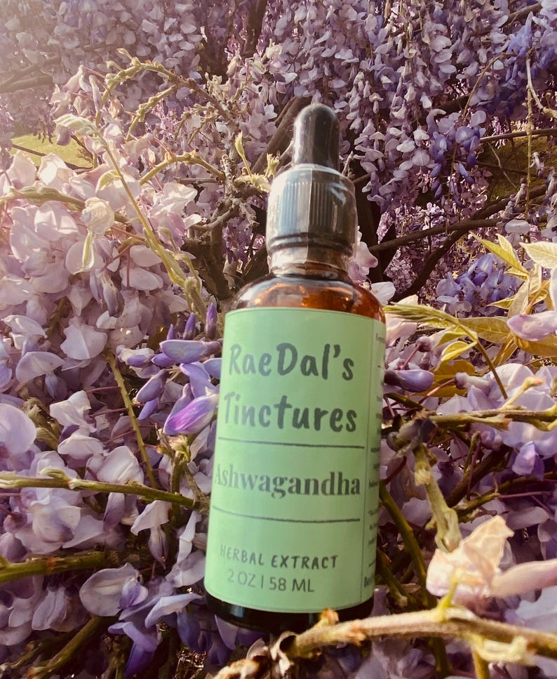 May include: A green glass bottle with a dropper top and a label that reads "RaeDal's Tinctures Ashwagandha Herbal Extract 2 oz | 58 ML". The bottle is sitting on a bed of purple flowers.
