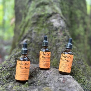 May include: Three amber glass bottles with black droppers, labelled "Rae Dal's Tinctures." The labels read "Burdock," "Ashwagandha," and "Valerian," with text indicating they are herbal extracts for dogs or cats. The bottles are on a mossy surface.
