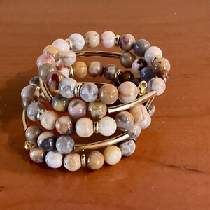 May include: A set of five bracelets made with brown, white, and gray agate beads. The bracelets are strung on gold-toned metal wire.