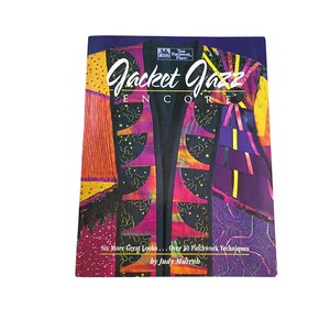 Op de afbeelding: Een softcover boek getiteld Jacket Jazz Encore door Judy Murrah met een kleurrijke patchwork jas op de omslag. Het ontwerp van de jas bevat zwarte, paarse, oranje en gele stoffen panelen met sterpatronen en verschillende texturen.
