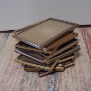 May include: A stack of rectangular picture frames with gold and silver-colored edges. The frames are stacked on top of each other, with a beige backing visible in some. The frames are on a multi-colored woven rug.