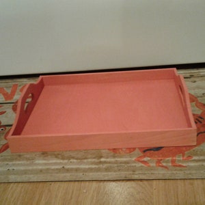 May include: A rectangular wooden serving tray with a coral pink finish and rounded edges. The tray has two handles on the sides.