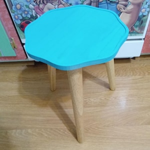 May include: A small, three-legged wooden stool with a bright turquoise, flower-shaped top. The legs are light-colored wood. The stool is on a wooden floor.