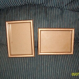 May include: Two empty picture frames with a wood-like finish. The frames are both rectangular and have a light brown color. The frames are sitting on a dark blue striped surface.