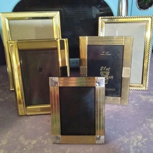 May include: A collection of gold-toned picture frames in various sizes. Some frames have a black backing, while others have a lighter backing. One frame has the text "21st Anniversary Frames". The frames are arranged on a purple surface.