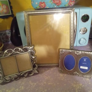 May include: Three silver-toned picture frames are displayed. One large rectangular frame stands vertically in the center. To the left is a smaller rectangular frame with ornate scrollwork. To the right is a horizontal frame with two oval openings.