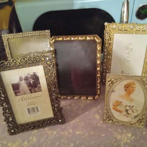 May include: A collection of four rectangular and oval picture frames in gold and bronze tones. The frames feature intricate metal filigree, embedded clear crystals, and decorative borders. One frame contains a photo of a couple, another a bride.