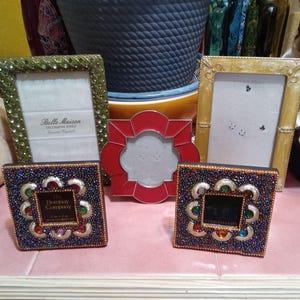 May include: A collection of decorative picture frames in various styles and colors. Includes a green frame, a gold frame, a red frame, and two square frames with jewel embellishments. The frames are empty.