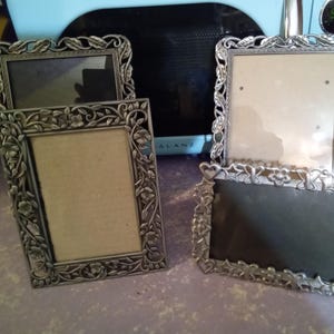 May include: Four ornate silver picture frames. Two frames have floral designs, and two have heart and star designs. The frames are rectangular and have a vintage aesthetic. The frames are empty, ready for photos.