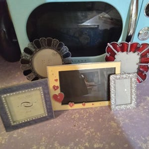 May include: A collection of decorative picture frames in various shapes and sizes. Frames include a round black frame, a rectangular gold frame with red hearts, a small silver frame, a blue square frame, and a red scalloped frame.