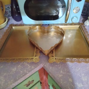May include: Gold-toned metal serving trays with ornate, filigree edges. The set includes two rectangular trays and a heart-shaped tray, all with a reflective surface. The trays are likely vintage or antique, suitable for decorative use.