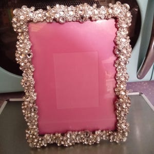 May include: A rectangular picture frame with a pink interior. The frame is embellished with numerous clear, sparkling rhinestones. The frame is silver-toned and has a decorative, ornate design. The background is a light blue color.