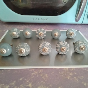 May include: A set of ten gray ceramic cabinet knobs with white floral patterns and gold-colored hardware. The knobs are arranged on a silver tray, with a light blue microwave in the background.