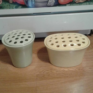 May include: Two cream-colored ceramic flower frogs. One is a tall, flared cylinder, and the other is an oval shape. Both have numerous holes in the top for arranging flowers. They sit on a wooden surface.