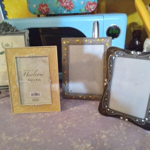 May include: Four decorative picture frames in various styles and colors. One gold frame has the text "Heirloom 4 x 6 in." The frames are silver, gold, and black, with ornate details. The frames are empty and ready for photos.
