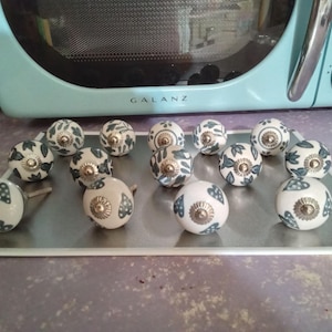 May include: A collection of 12 white ceramic drawer knobs with a blue and green floral design. The knobs have a silver accent in the center.
