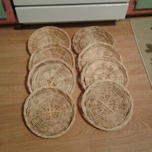 May include: Ten round woven wicker coasters in a natural beige color. The coasters are stacked in a pile on a wooden floor.