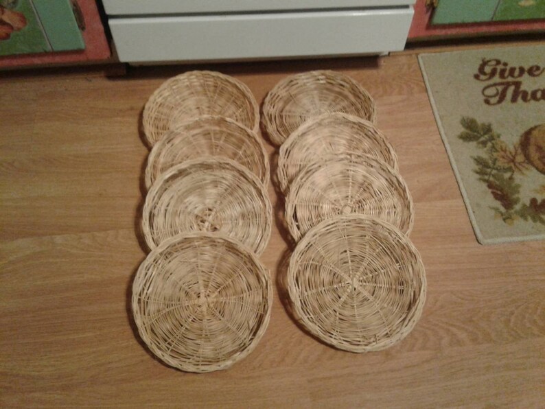 May include: Ten round woven wicker coasters in a natural beige color. The coasters are stacked in a pile on a wooden floor.