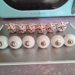 May include: A collection of 12 white ceramic knobs with a crackled finish and silver accents. The knobs have a floral design in shades of orange, red, and purple.