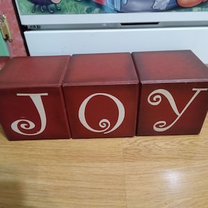 May include: Three red wooden blocks spelling the word "JOY" in white, decorative lettering. The blocks are arranged side-by-side on a wooden surface. The letters have a whimsical, swirly design.