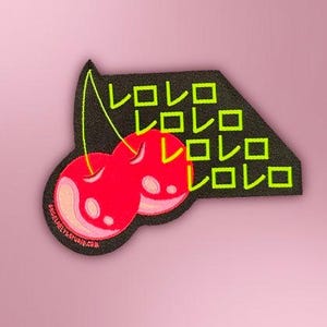May include: A black embroidered patch featuring two bright red cherries with light pink highlights and a lime green stem. The patch includes the Japanese katakana text "レロレロ" repeated four times in lime green, with the text "ANGELBELLYSTUDIO.COM" printed in small white letters along the bottom edge.