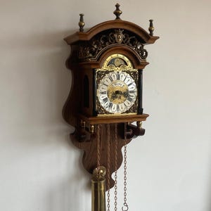 Peut inclure: Une horloge murale en bois au design décoratif. Le cadran est doré et blanc avec des chiffres romains noirs. Deux poids en laiton sont suspendus en dessous, reliés par des chaînes. L'horloge est fixée sur un support en bois.